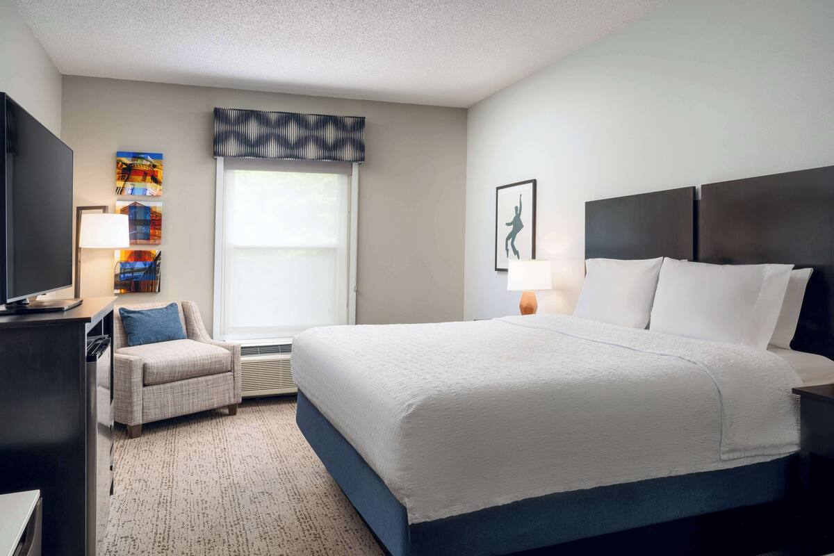 A hotel room with a large bed, a desk with a computer, a chair, a TV, and a window with blinds.