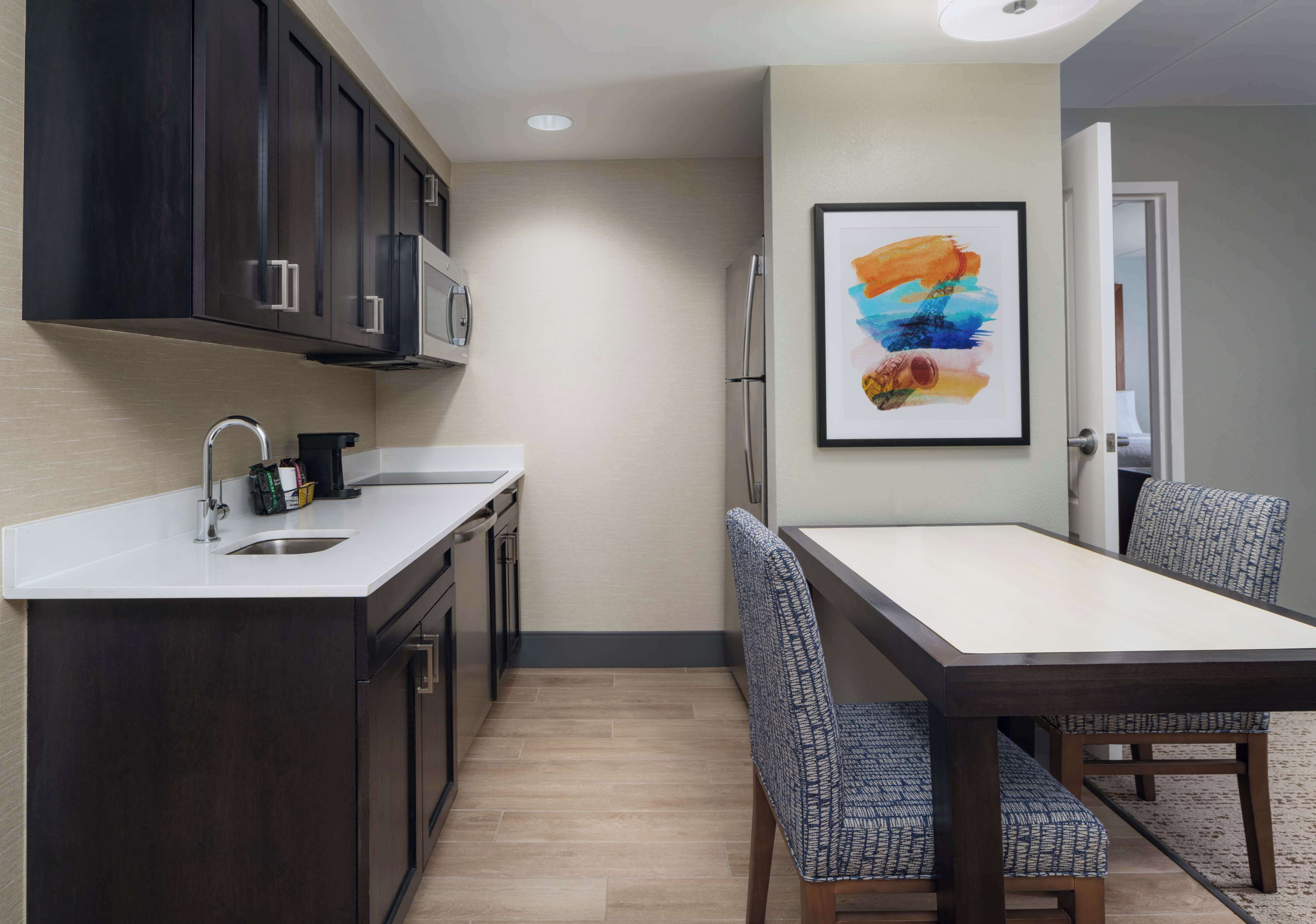 A modern kitchen with dark cabinets, a white countertop, and a dining area with a table and patterned chairs.