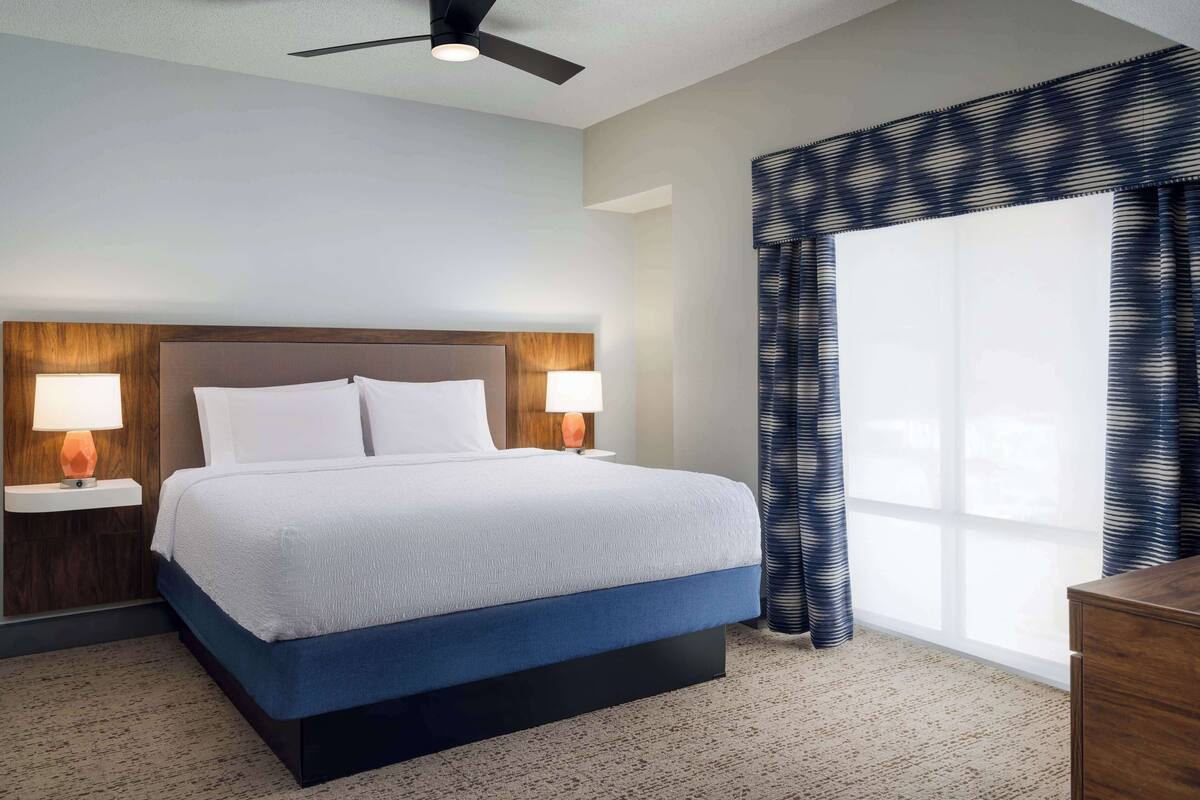 A hotel room with a bed, two bedside lamps, a ceiling fan, and a large window with curtains.