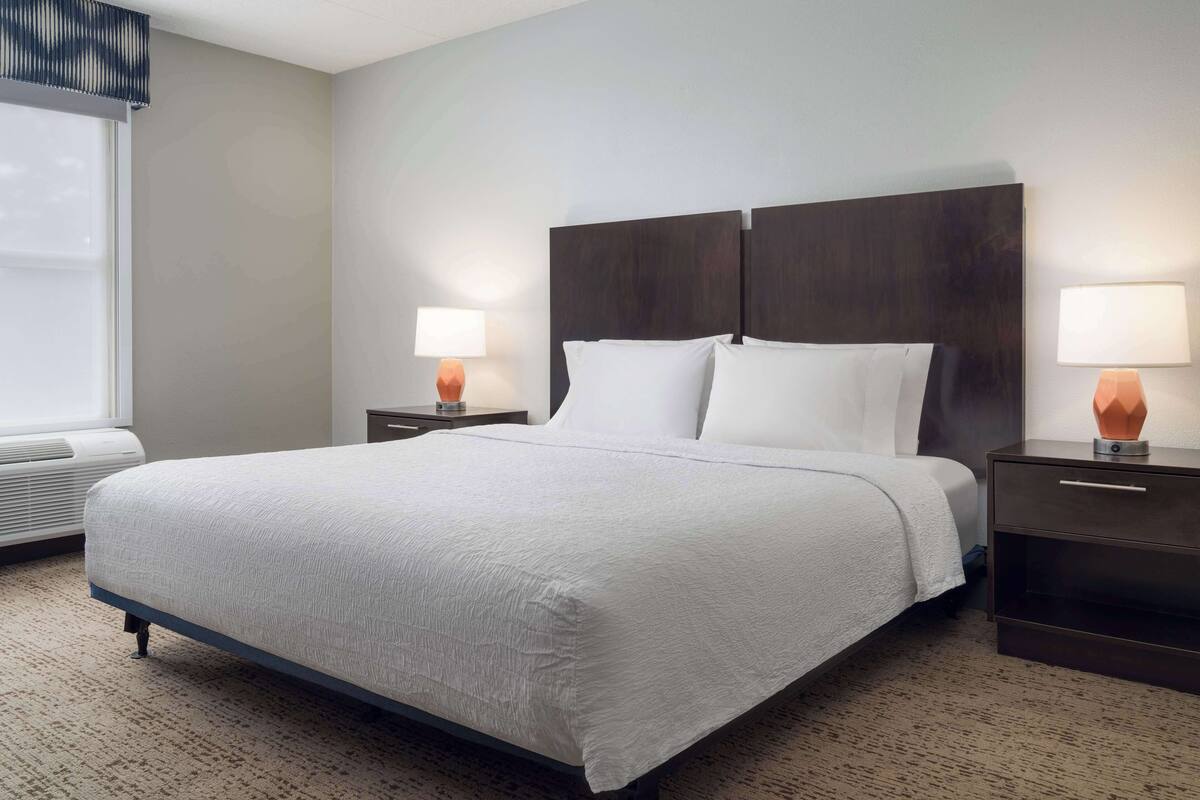 A neatly made bed with a dark headboard, two bedside tables with lamps, and a window with blinds.