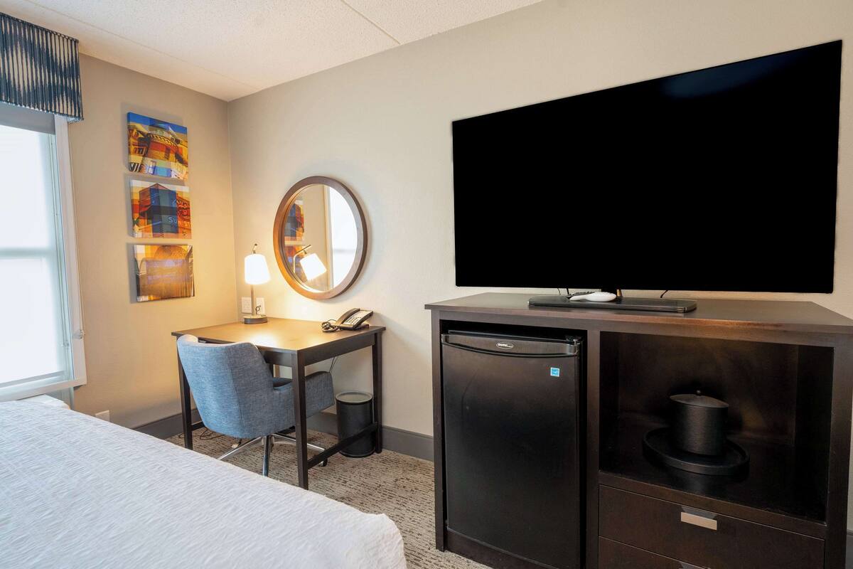 A hotel room with a bed, a desk with a chair, a television, a refrigerator, and a trash can.