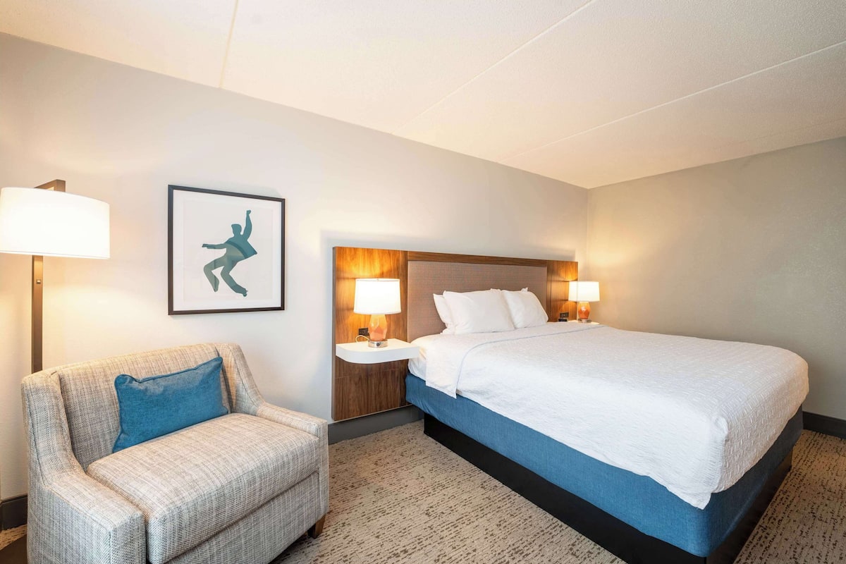 A hotel room with a bed, a chair, two bedside tables with lamps, and a framed picture on the wall.