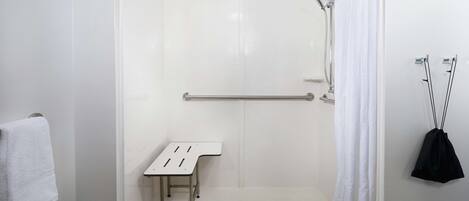 Una ducha sin borde con pared de azulejos blancos, una barra de apoyo, una mesita y un perchero.