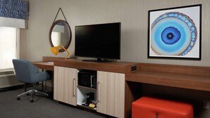 A hotel room with a wooden desk, a flat-screen TV, a blue office chair, a red suitcase, a framed abstract painting, and a wall-mounted mirror. - Hampton Inn Niagara Falls (Niagara Falls)