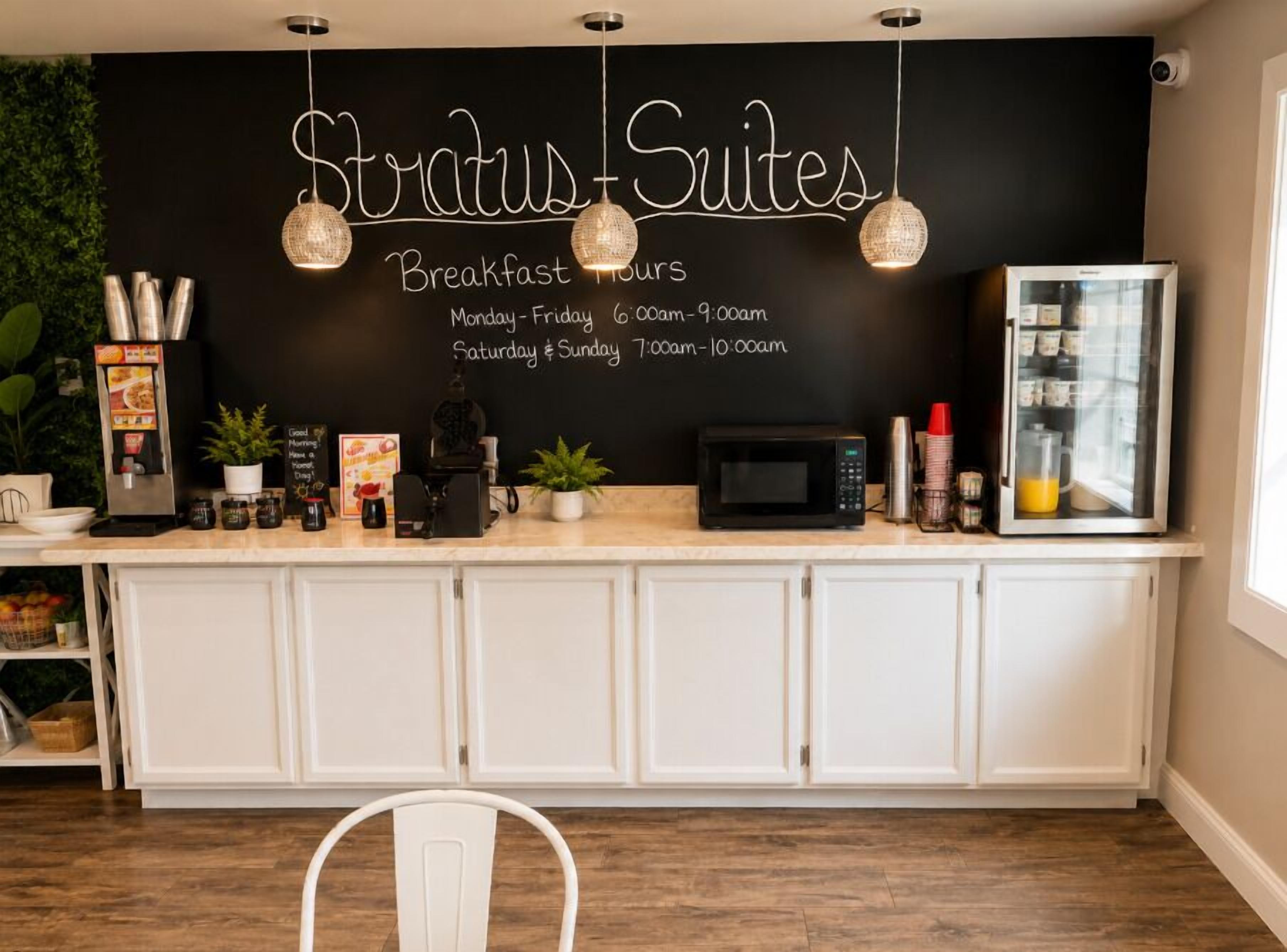 A kitchen area with a chalkboard displaying breakfast hours, a coffee machine, a microwave, and a refrigerator.