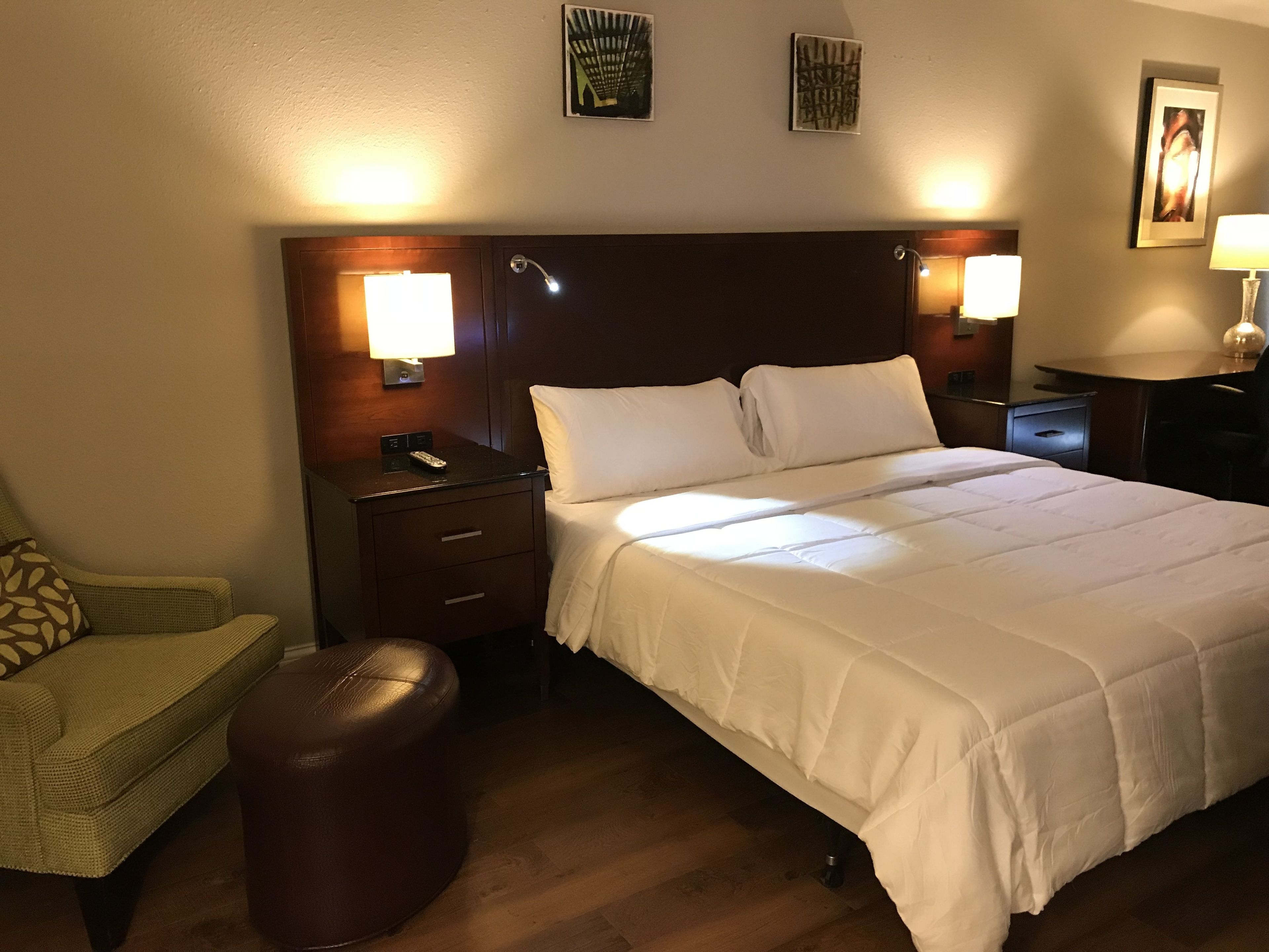 A hotel room with a large bed, a nightstand with a lamp, a chair, and a small ottoman.