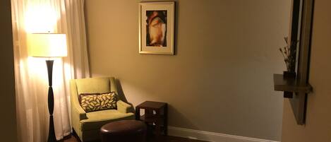 A room with a green armchair, a small table, a lamp, and a painting on the wall.