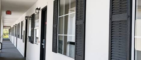 Un couloir d’hôtel avec des volets noirs aux fenêtres, une façade blanche et un panneau indiquant la sortie.