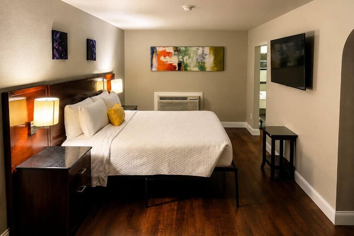 A hotel room with a bed, bedside lamps, a flat-screen TV, a painting on the wall, and a heater.