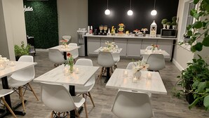 Un comedor moderno con mesas y sillas blancas, una encimera de cocina con diferentes alimentos y una pared verde con un letrero.