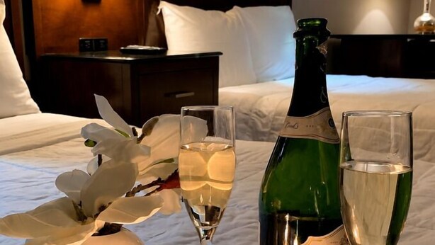 Habitación de hotel con cama, mesita de noche con lámpara, una botella de champán, dos copas, un florero con flores y dos velas encendidas sobre una bandeja.