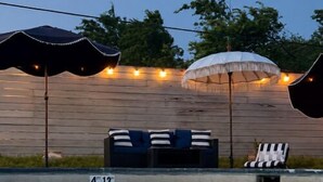 Una piscina con sillones de descanso y sombrillas, rodeada por una terraza de madera y luces colgantes.