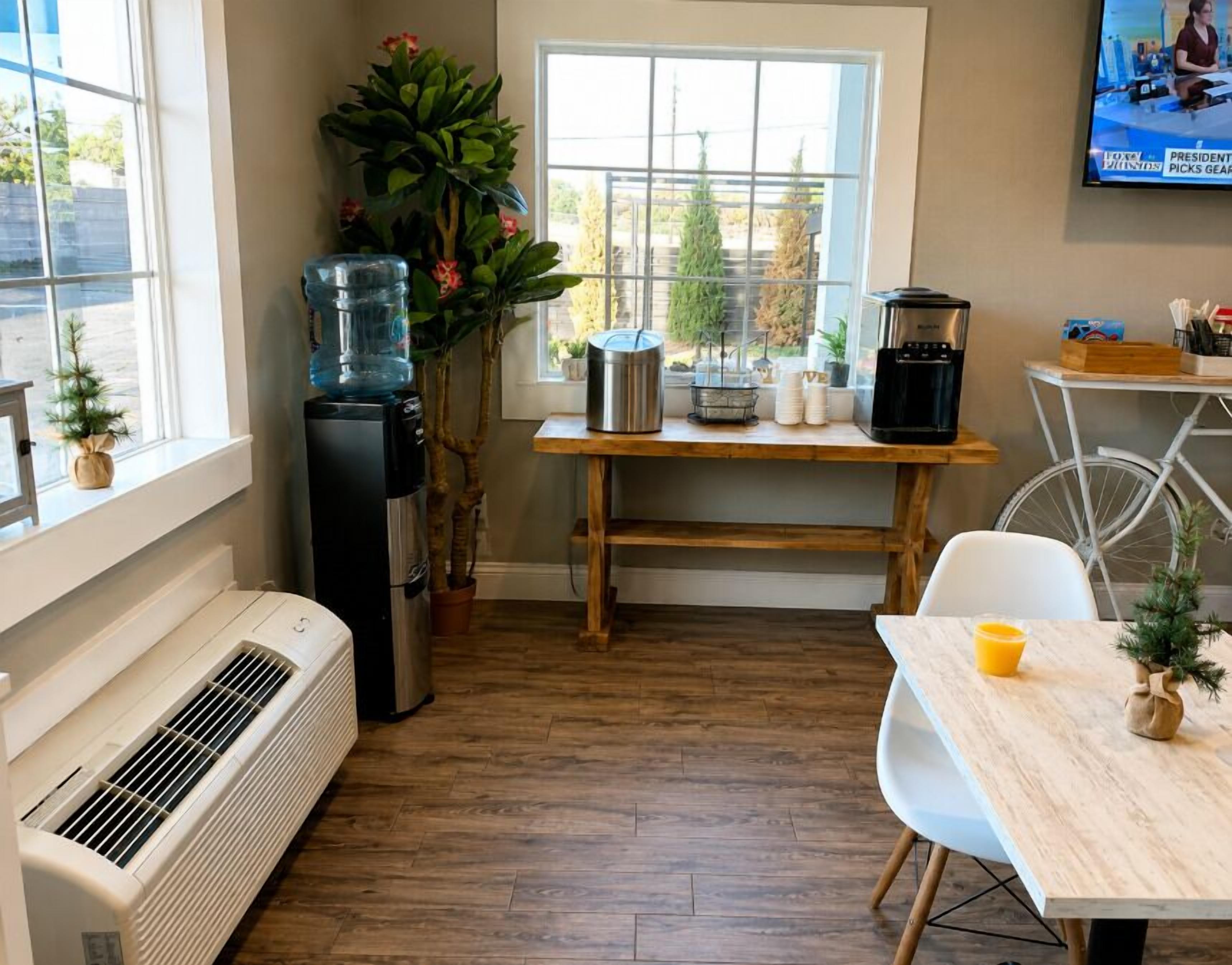 Una cocina con dispensador de agua, cafetera, refrigerador pequeño, mesa de madera y una silla.