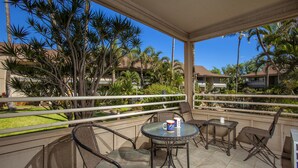 A covered outdoor area with wicker furniture and a table, surrounded by palm trees and a view of other buildings.