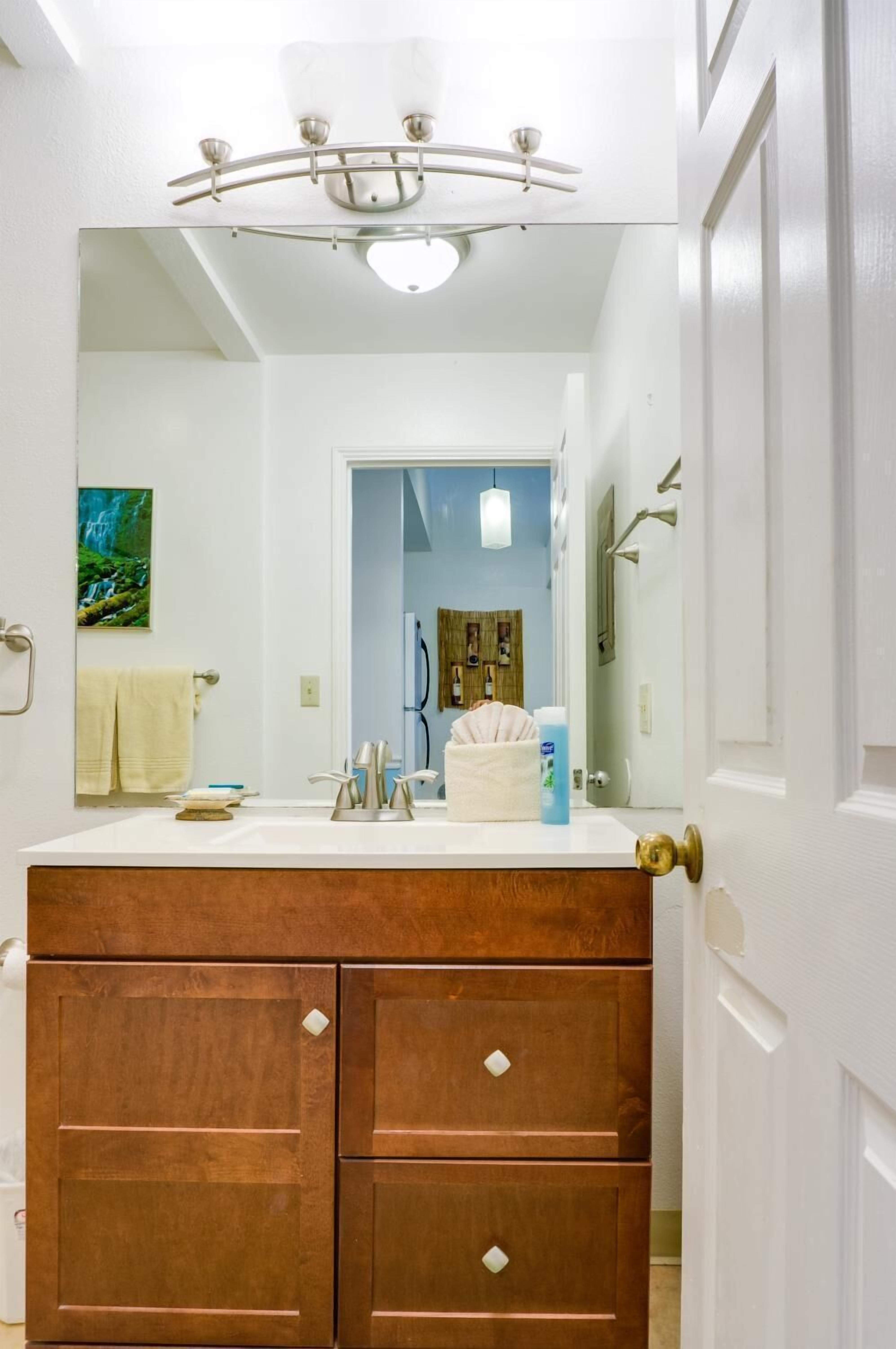 A bathroom with a large mirror, a wooden vanity with drawers, a sink, and a shower area visible through the doorway.