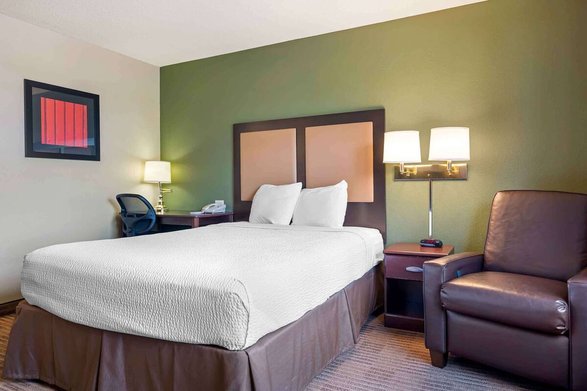 Een hotelkamer met een groot bed, een bureau met stoel, een bruine leren fauteuil en twee bedlampjes.