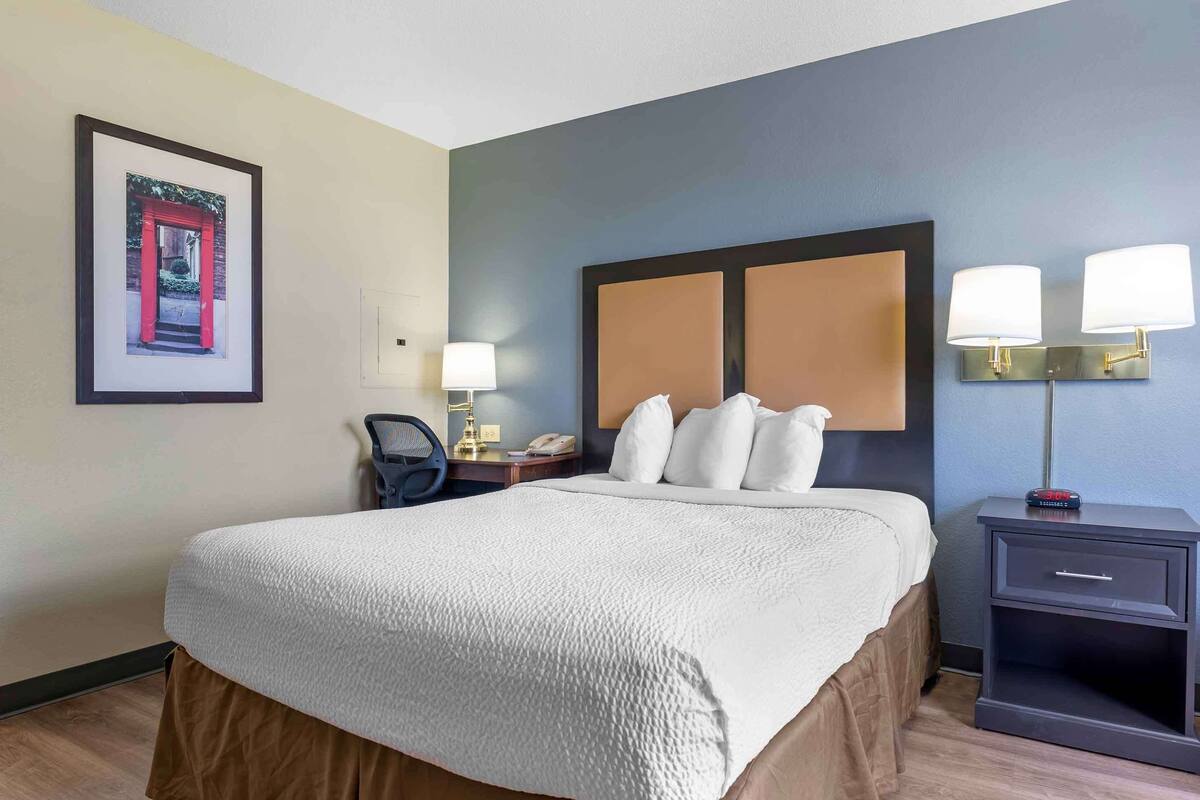 Een hotelkamer met een groot bed, een nachtkastje met een lamp, een lijst met een foto aan de muur en een stoel.