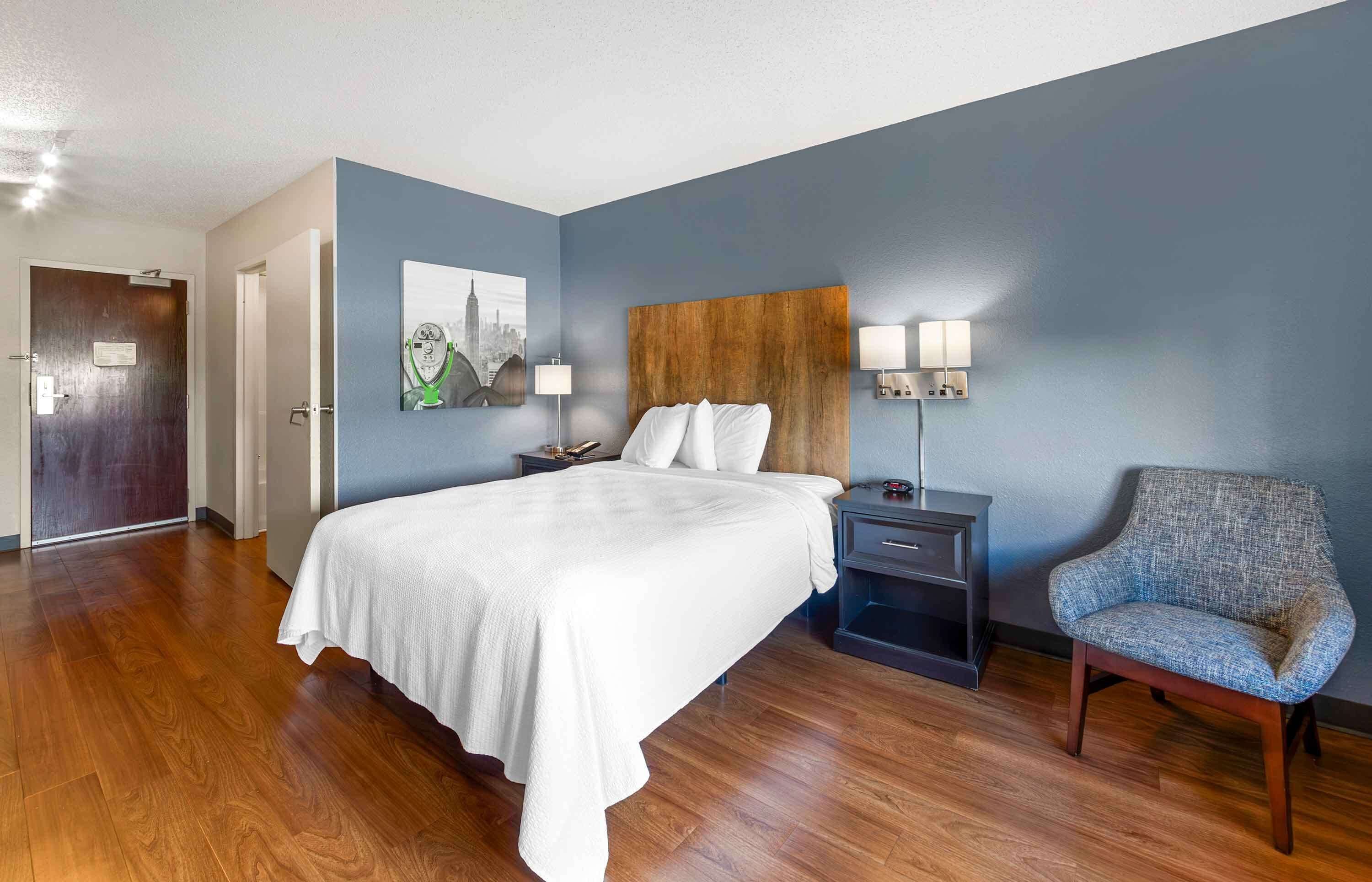 Une chambre d’hôtel avec un grand lit, une table de chevet avec des lampes, une chaise et un tableau représentant un paysage urbain accroché au mur.
