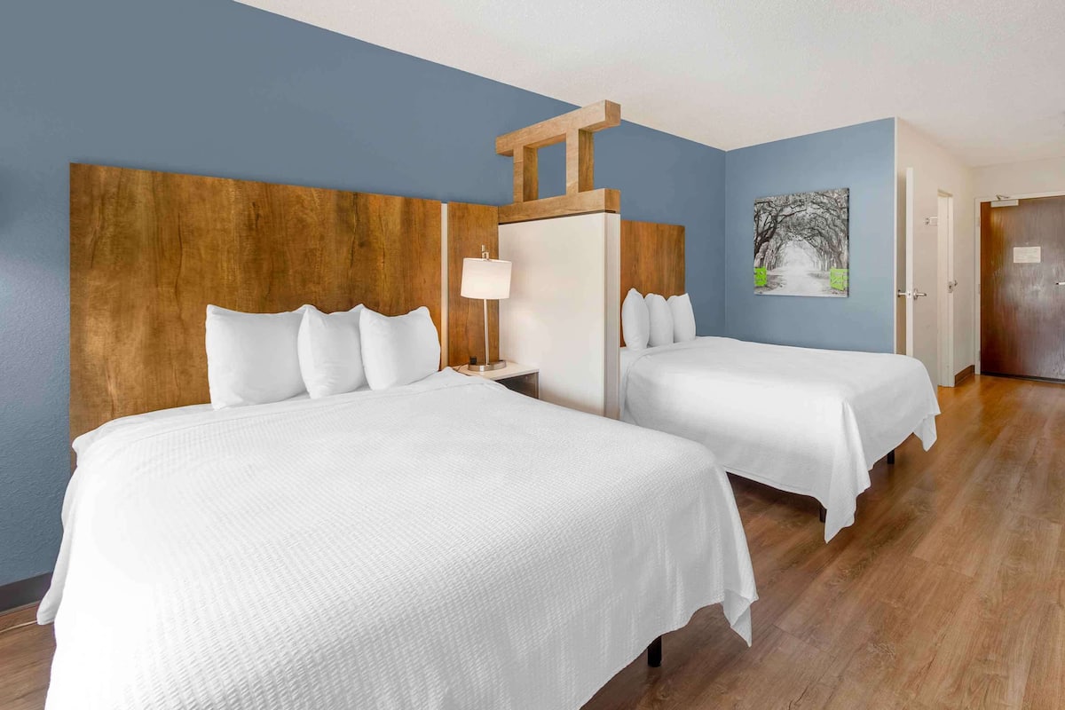 Habitación de hotel con dos camas, cabecera de madera, mesita de noche con lámpara y un cuadro en la pared.