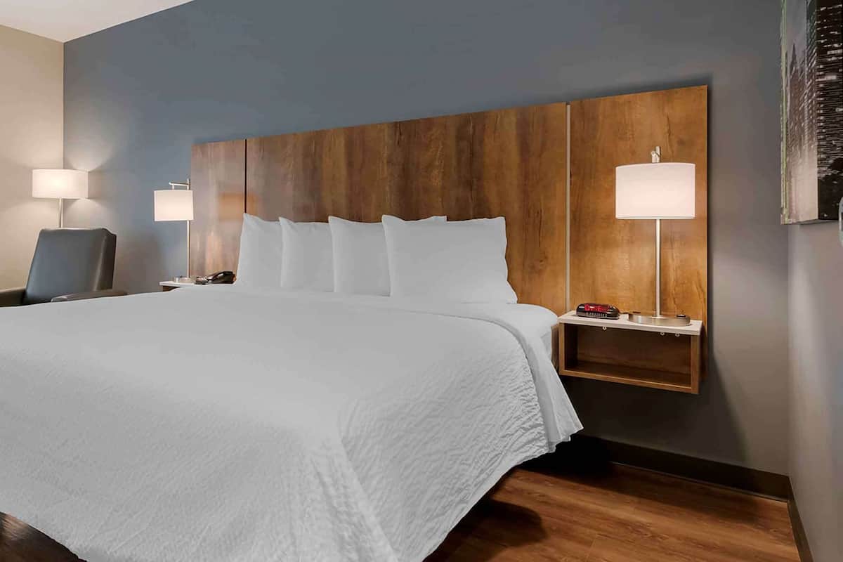 Habitación de hotel con una cama grande, cabecera de madera, dos lámparas de noche, un escritorio con lámpara y una ventana con persianas.