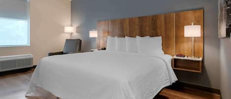 A hotel room with a large bed, a wooden headboard, two bedside lamps, a desk with a lamp, and a window with blinds.