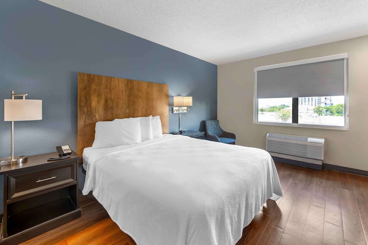 Habitación de hotel con una cama grande, una mesita de noche con lámpara, una silla, ventana con vistas y un equipo de aire acondicionado.