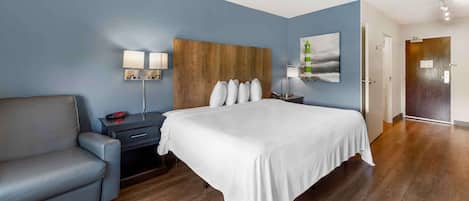Quarto de hotel com uma cama grande, um sofá cinzento, um painel de cabeceira de madeira e um quadro na parede.