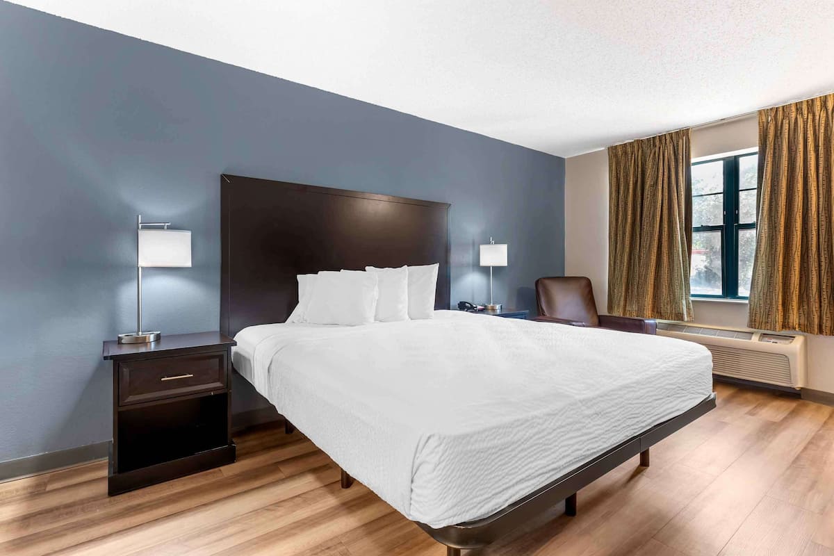 Ein modernes Hotelzimmer mit einem großen Bett, einem dunklen Holz-Kopfteil, zwei Nachttischen mit Lampen, einem Sessel und einem Fenster mit Vorhängen.