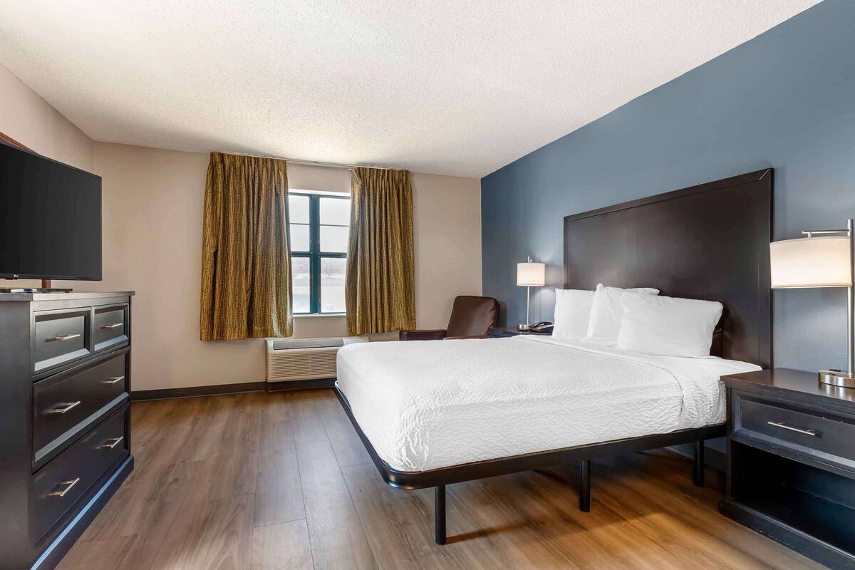Ein modernes Hotelzimmer mit einem großen Bett, einem Schreibtisch, einem Sessel, einem Fernseher und einem Fenster mit Vorhängen.