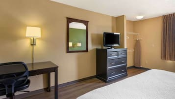 A hotel room with a bed, a desk with a chair, a television, a dresser, a mirror, and a lamp.