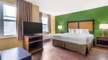 A hotel room with a large bed, a television on a wooden stand, a bedside table with a lamp, and a window with curtains.