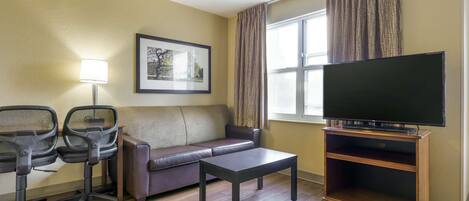 Une chambre d’hôtel avec un canapé, deux fauteuils de bureau, une table basse, un téléviseur posé sur un meuble en bois, un tableau encadré au mur et une fenêtre avec des rideaux.