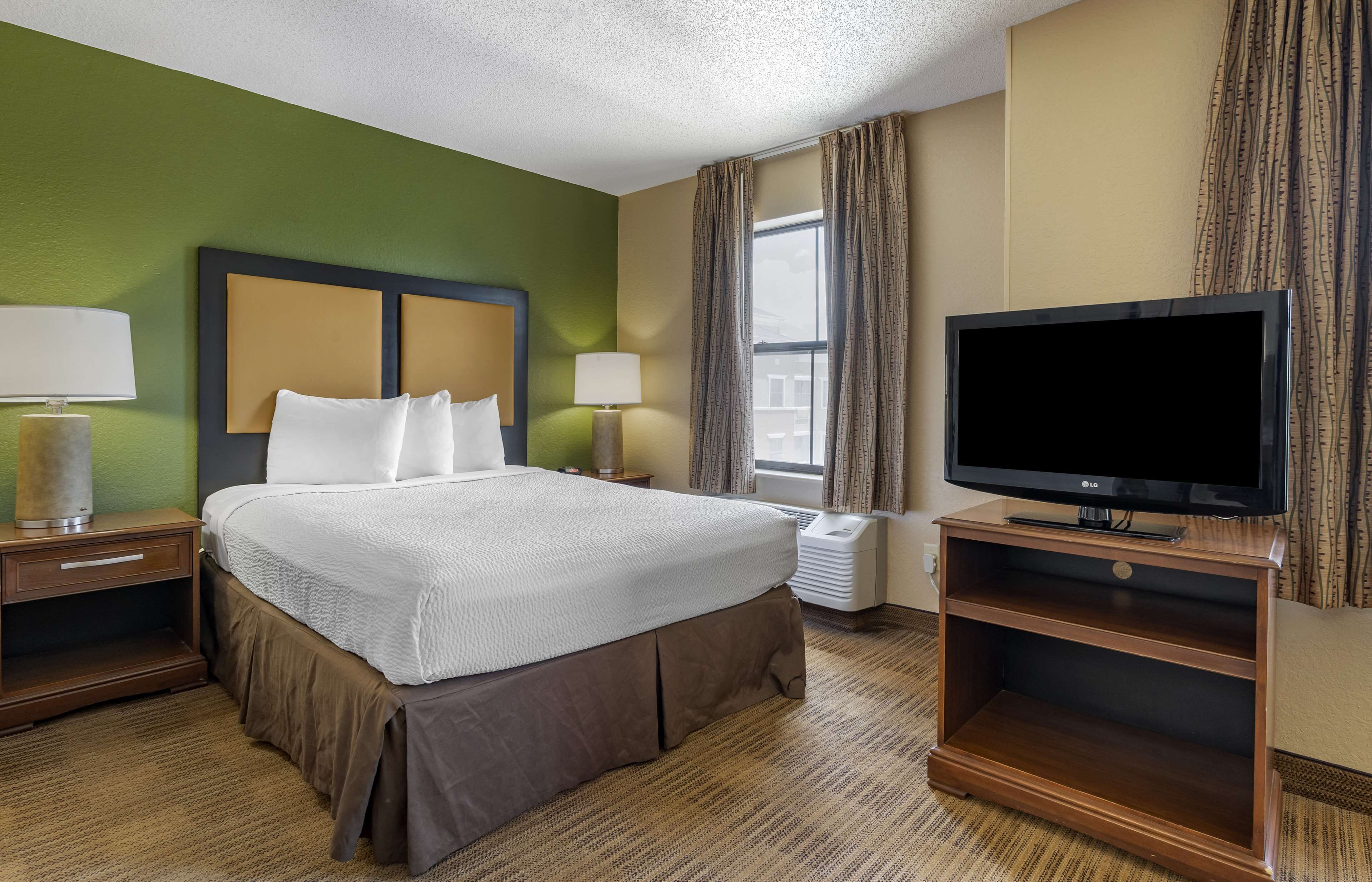 A hotel room with a large bed, two bedside tables with lamps, a flat-screen TV on a wooden stand, and a window with curtains.