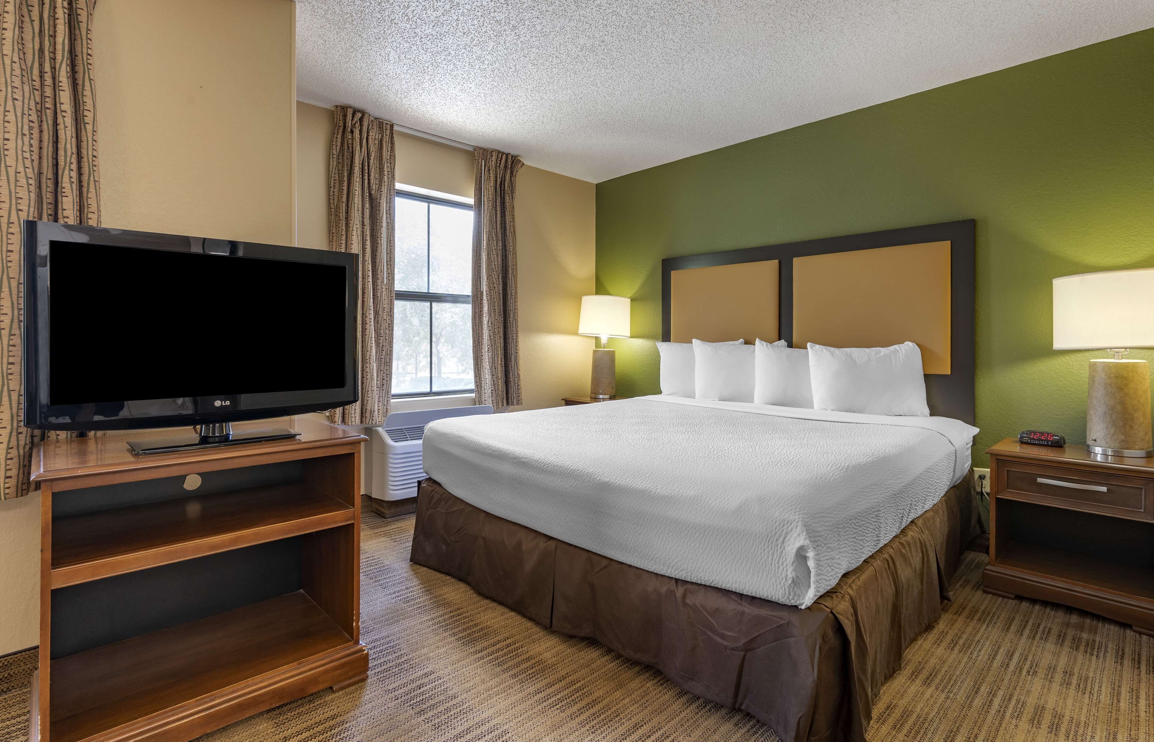 Habitación de hotel con una cama grande, un televisor de pantalla plana sobre un soporte de madera, una mesita de noche con lámpara y una ventana con cortinas.