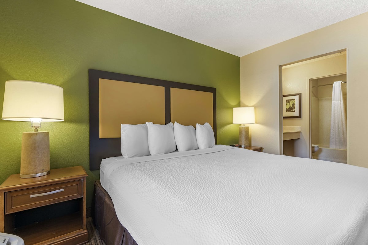 A hotel room with a large bed, two bedside lamps, a nightstand with a lamp, and a bathroom visible through an open door.