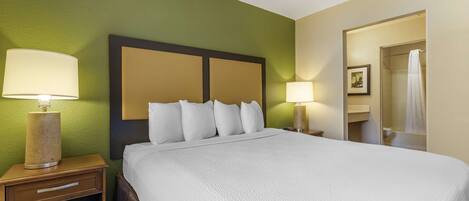 A hotel room with a large bed, two bedside lamps, a nightstand with a lamp, and a bathroom visible through an open door.