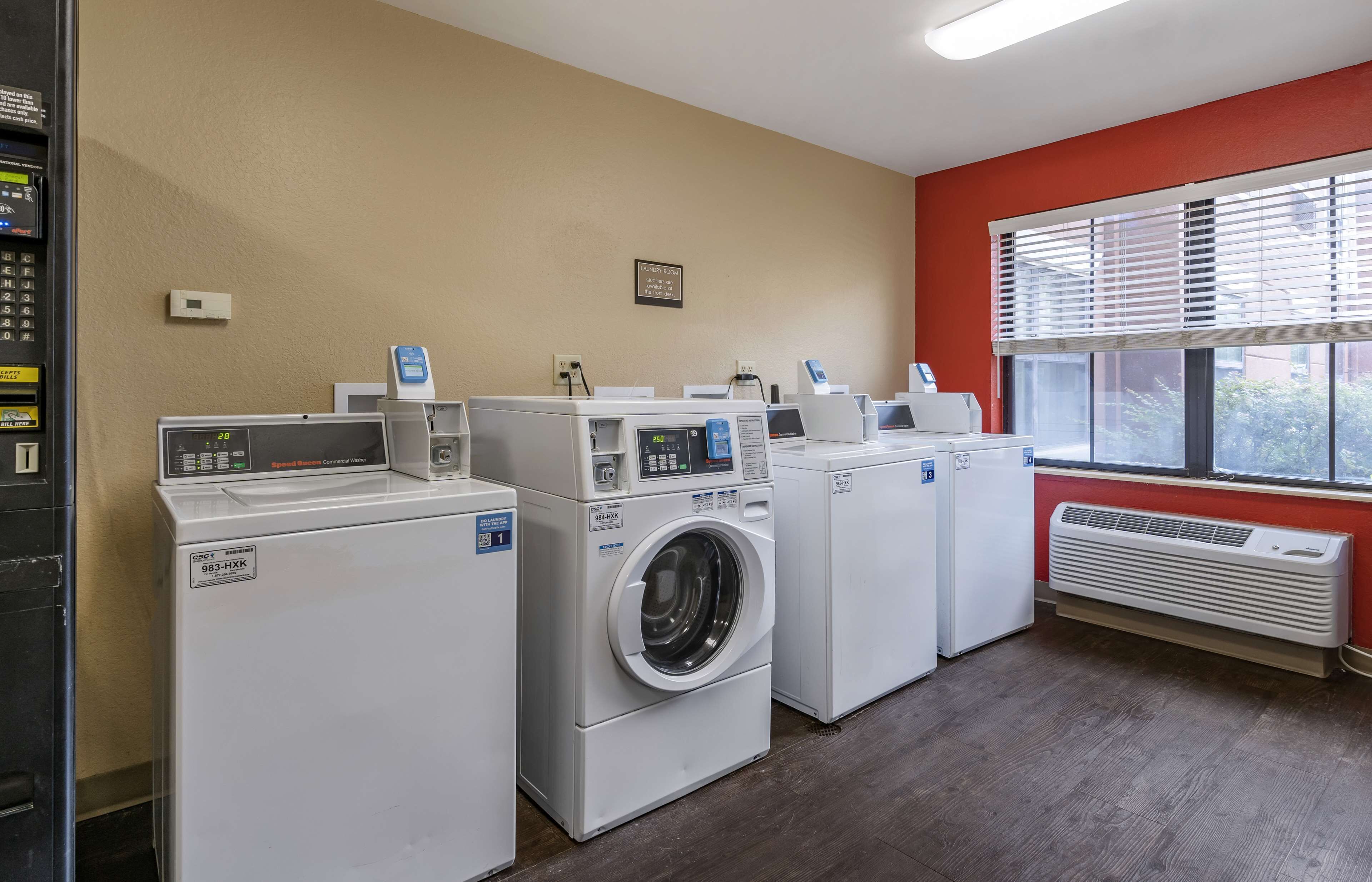 Une buanderie équipée de plusieurs machines à laver et de plusieurs sèche-linge, d’un panneau de commande et d’une fenêtre avec des stores.