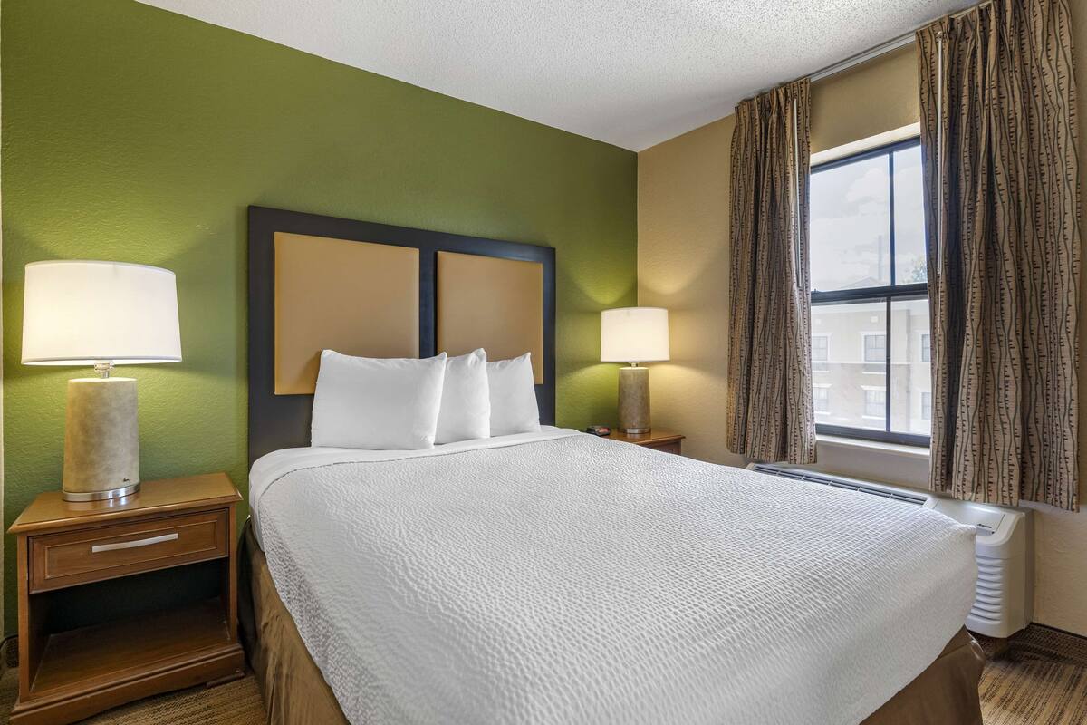 A hotel room with a large bed, two bedside lamps, a nightstand with a drawer, and a window with curtains.