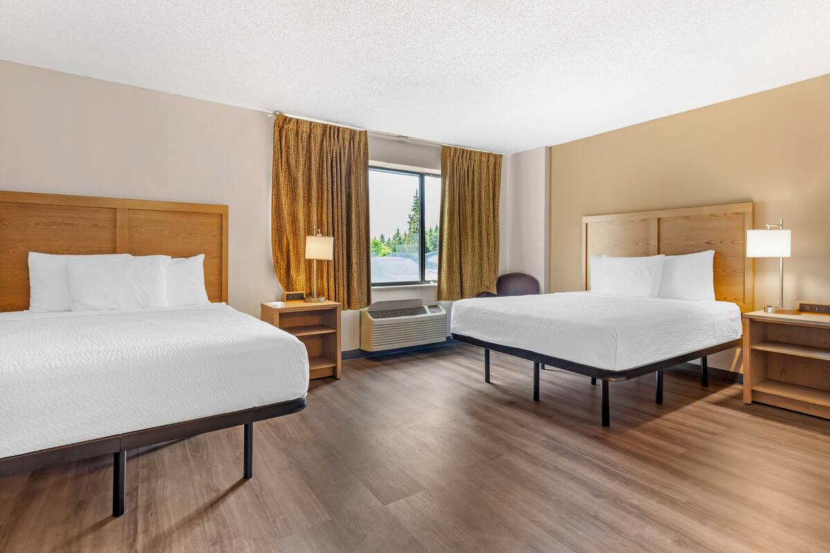 Hotellihuone, jossa on kaksi sänkyä, puiset päätyosat, ikkuna verhoilla, työpöytä, tuoli ja ilmastointilaite.