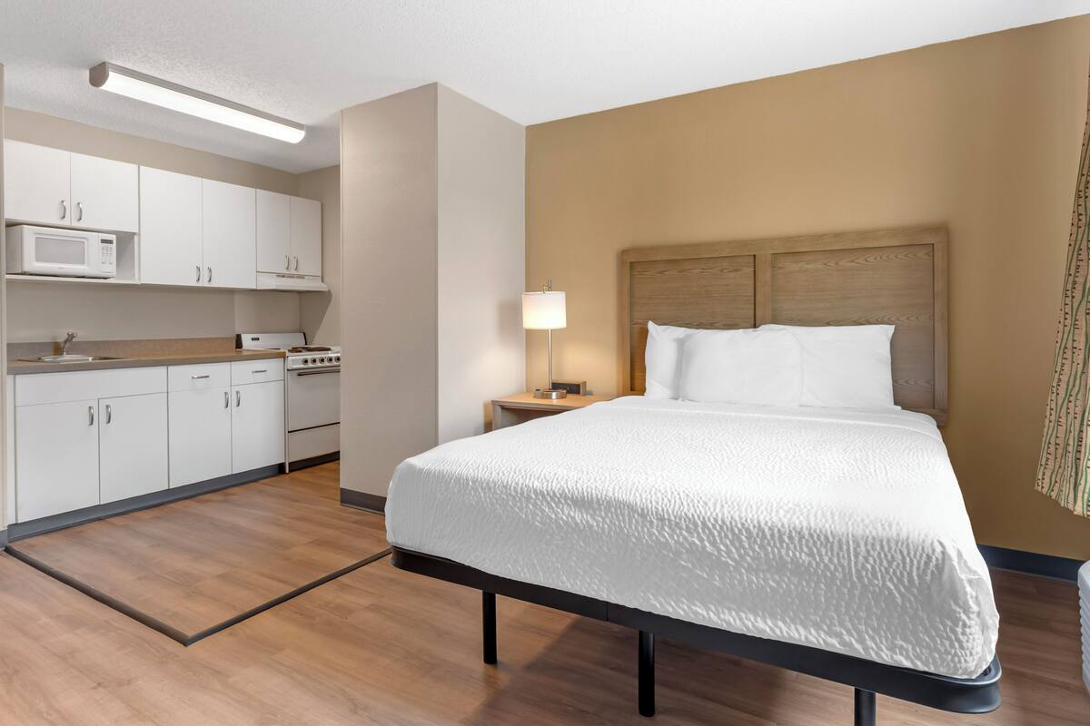 Hotellihuone, jossa on sänky, yöpöytä, lamppu, mikroaaltouuni ja pieni keittiö.