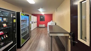 Un couloir étroit où l’on trouve des distributeurs automatiques, une rangée de casiers blancs et une longue table.