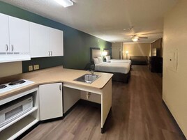 A compact hotel room with a kitchenette, a bed, a bedside table, and a ceiling fan.