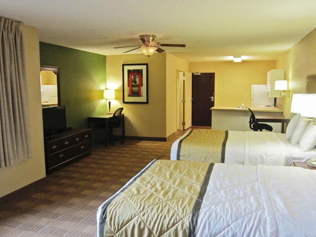 A hotel room with two beds, a desk, a chair, a ceiling fan, and a door.