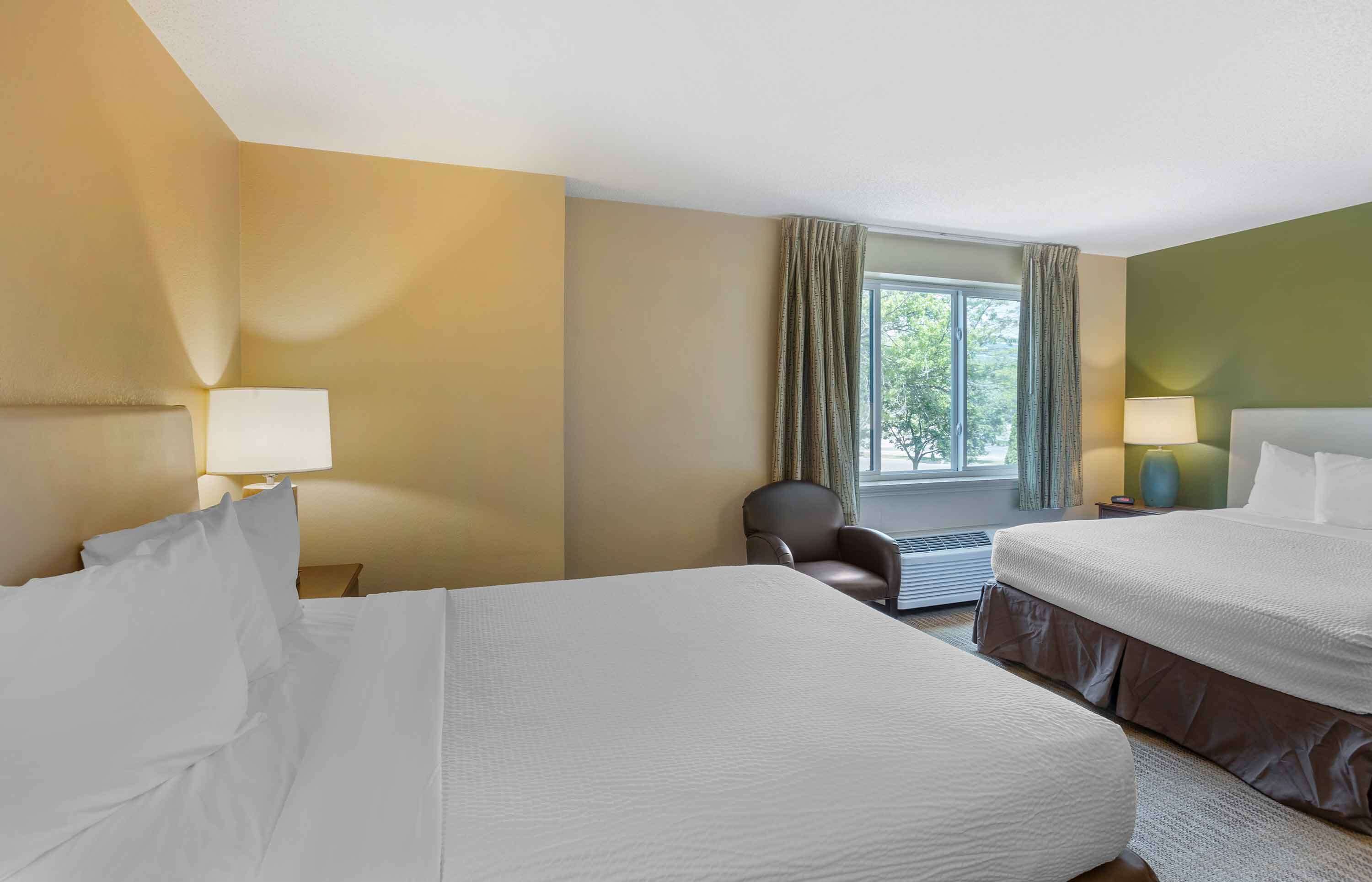 A hotel room with two beds, a chair, a window with curtains, and two bedside lamps.