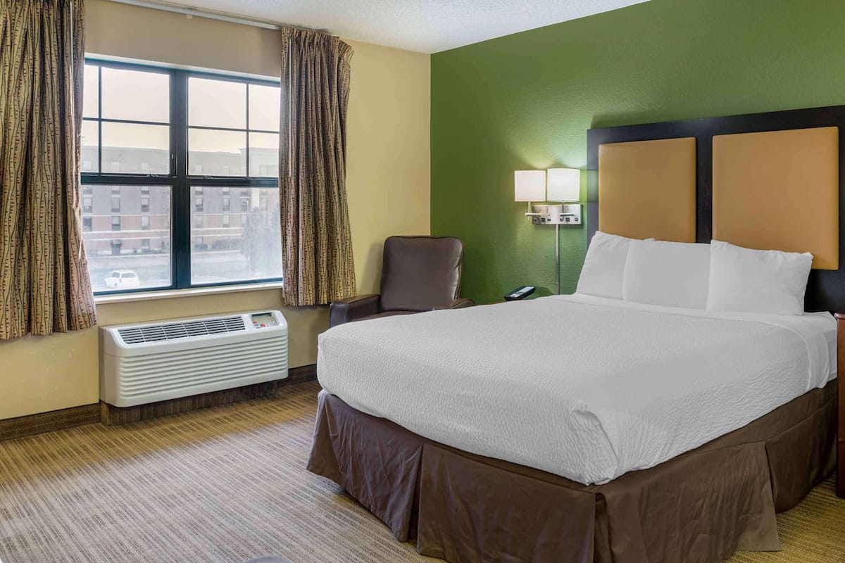 A hotel room with a large bed, a chair, a desk, a lamp, and a window with curtains.