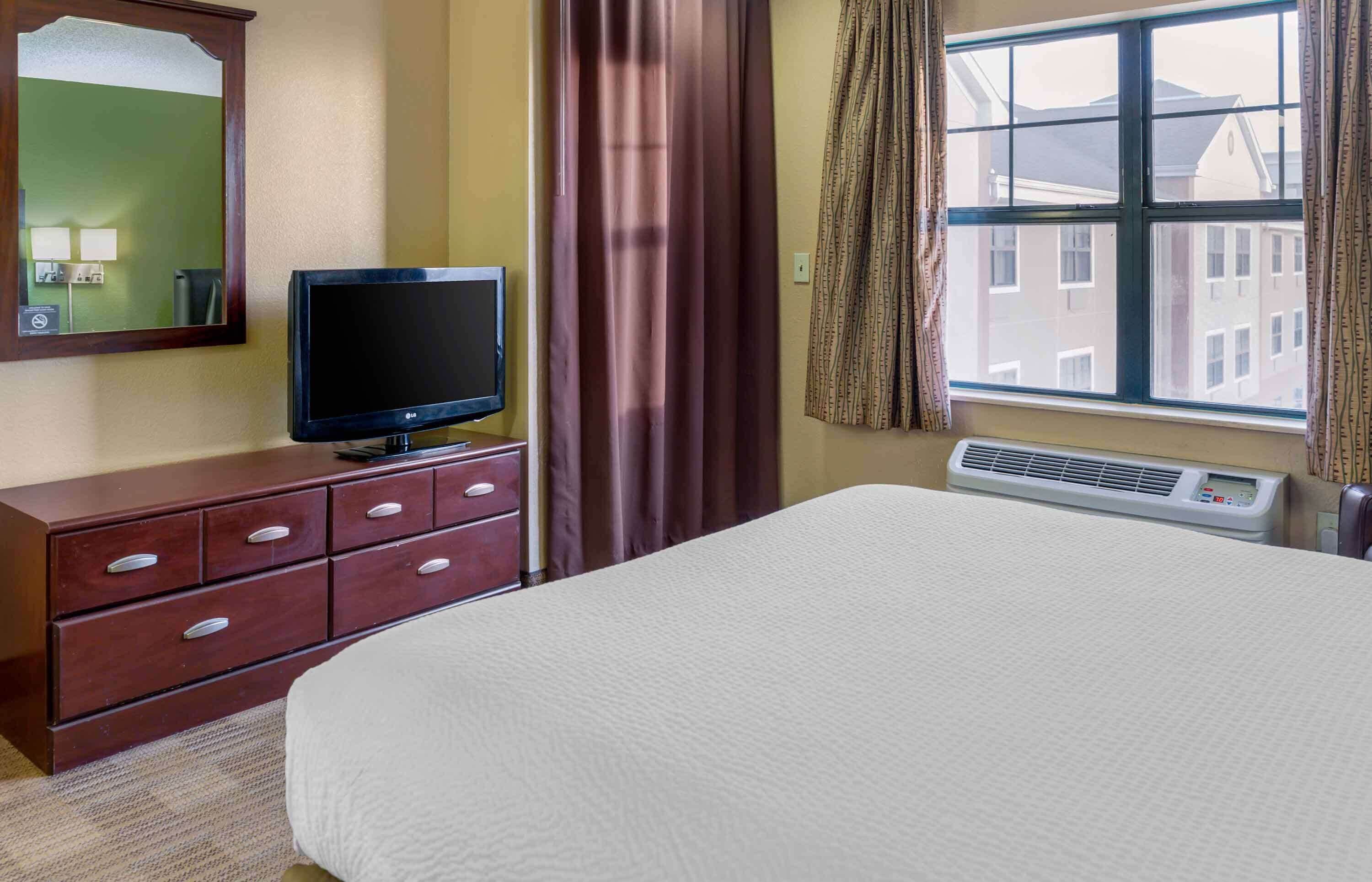 A hotel room with a bed, a TV on a wooden dresser, a window with curtains, and an air conditioning unit.