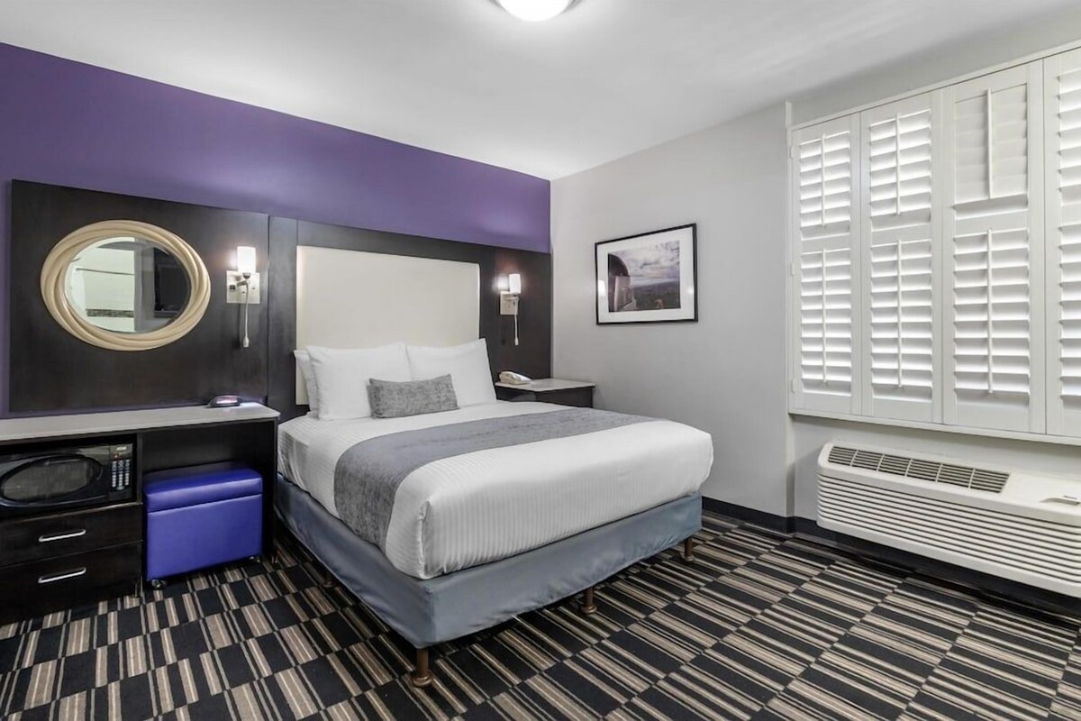 Ein Hotelzimmer mit Bett, Schreibtisch mit Computer, Mikrowelle, blauem Aufbewahrungsbehälter, großem Spiegel, Bild an der Wand und Fenster mit Jalousien.
