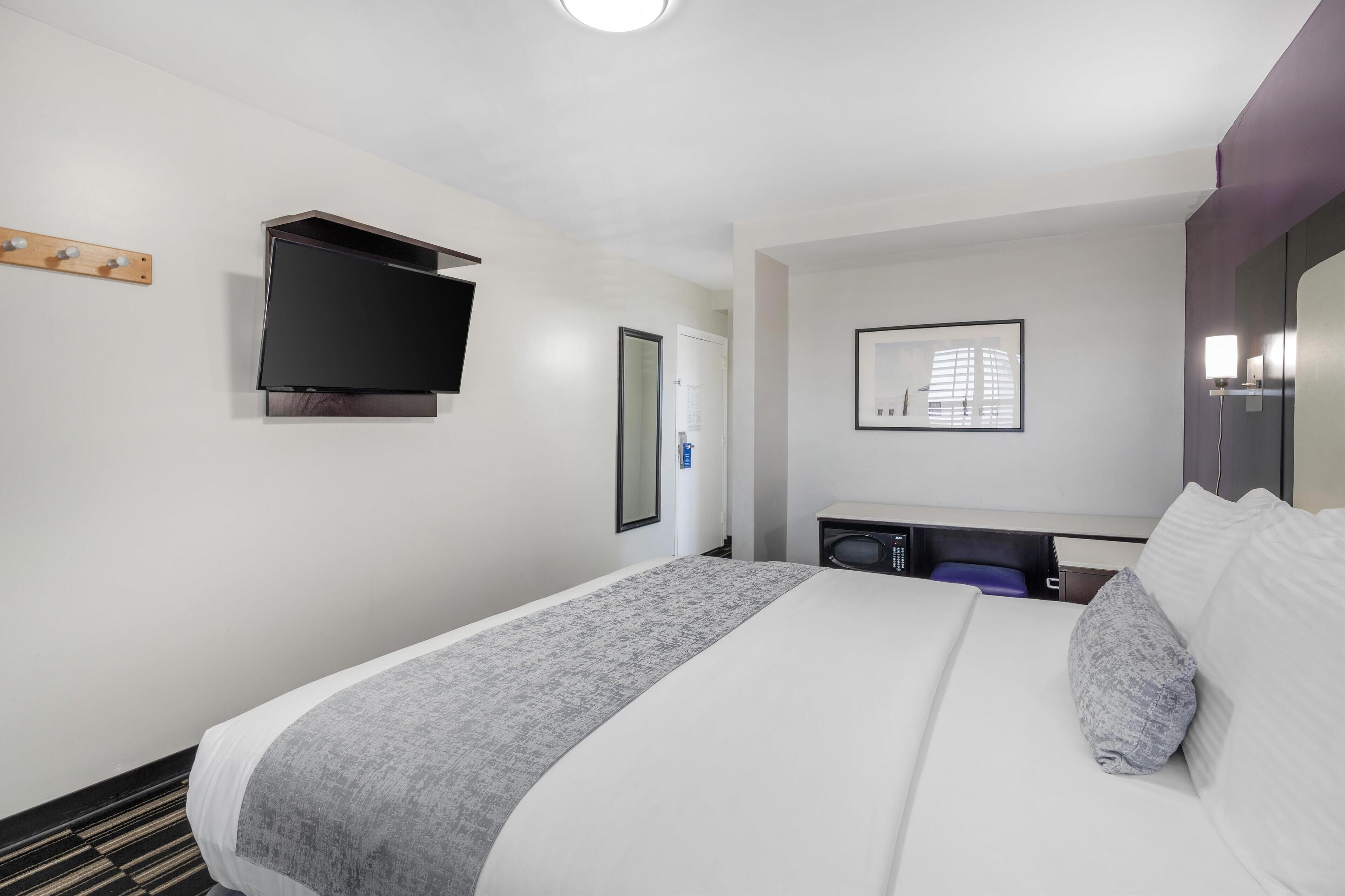 Habitación de hotel con cama, televisor de pantalla plana en la pared, escritorio pequeño con silla y un cuadro enmarcado.