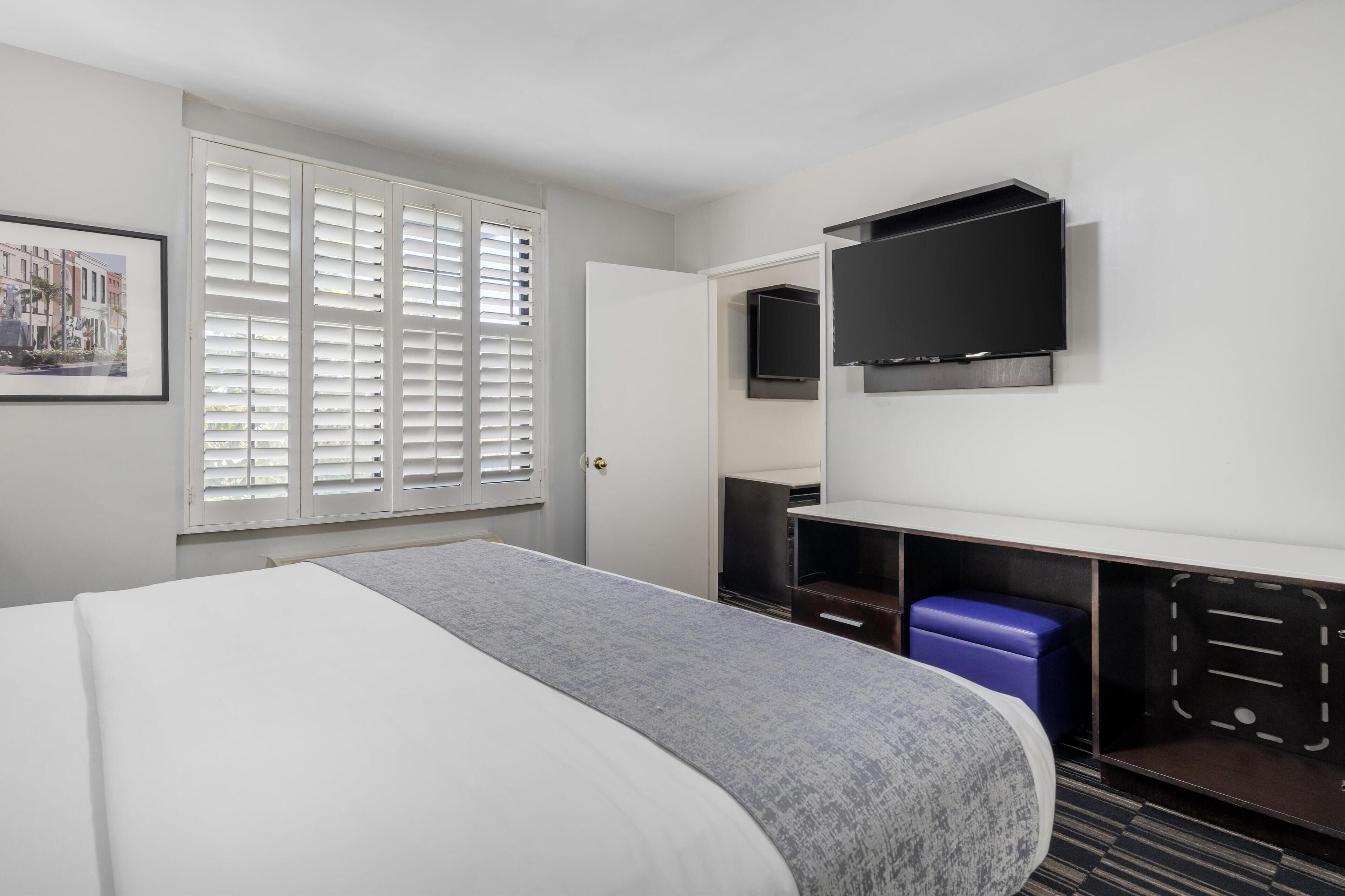 Una habitación de hotel con cama, escritorio con computadora, televisión y ventana con persianas.