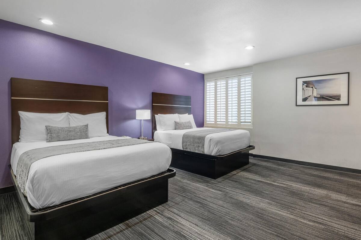 A hotel room with two beds, a window with blinds, a framed picture on the wall, and a carpeted floor.